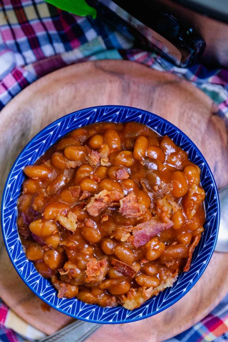 Overhead view of baked beans with crispy bacon pieces in blue-rimmed bowl on wooden plate, checkered cloth background, Easy Slow Cooker Dinn