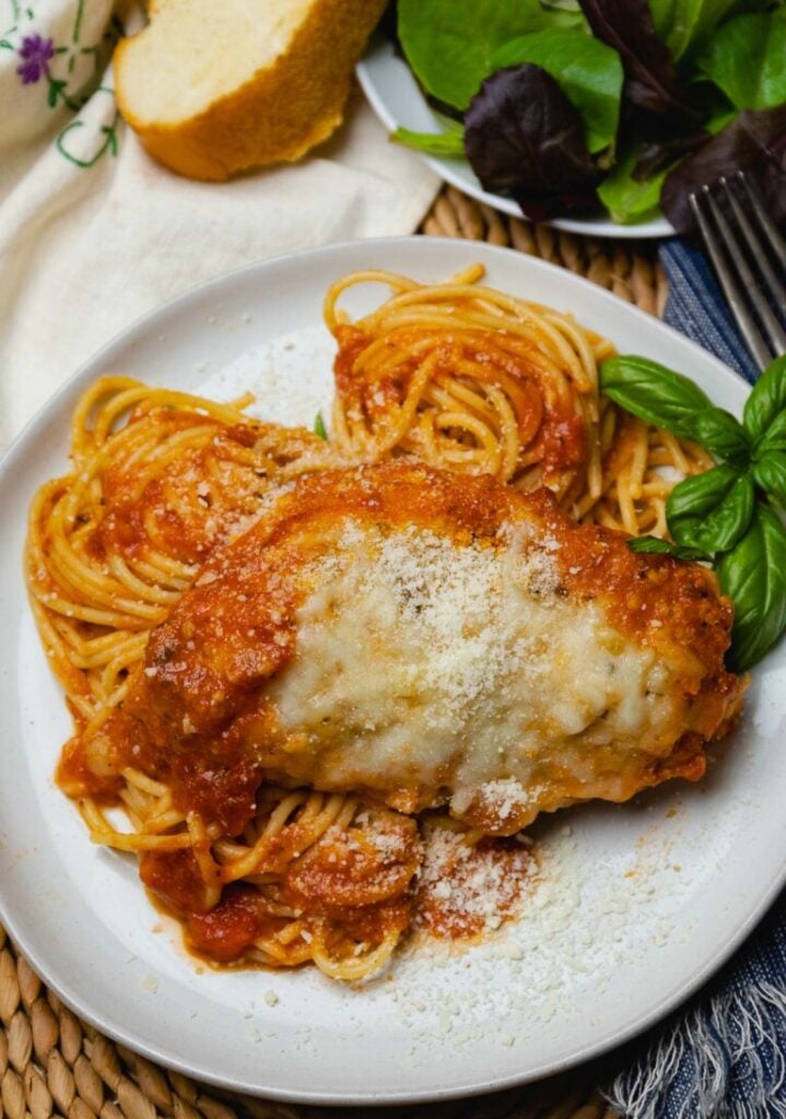 Chicken parmesan over spaghetti with melted cheese on white plate, fresh basil garnish, bread and salad visible, Easy Slow Cooker Dinners fo