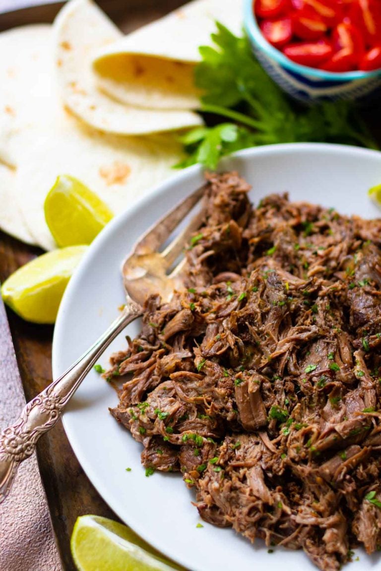 Shredded beef carnitas on white plate with lime wedges, tortillas, cilantro, and red peppers for Easy Slow Cooker Dinners for Busy Weeknight