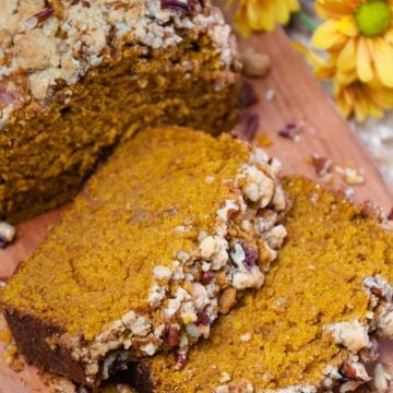 Sliced pumpkin pecan quick bread with crumb topping on wooden board beside yellow flowers