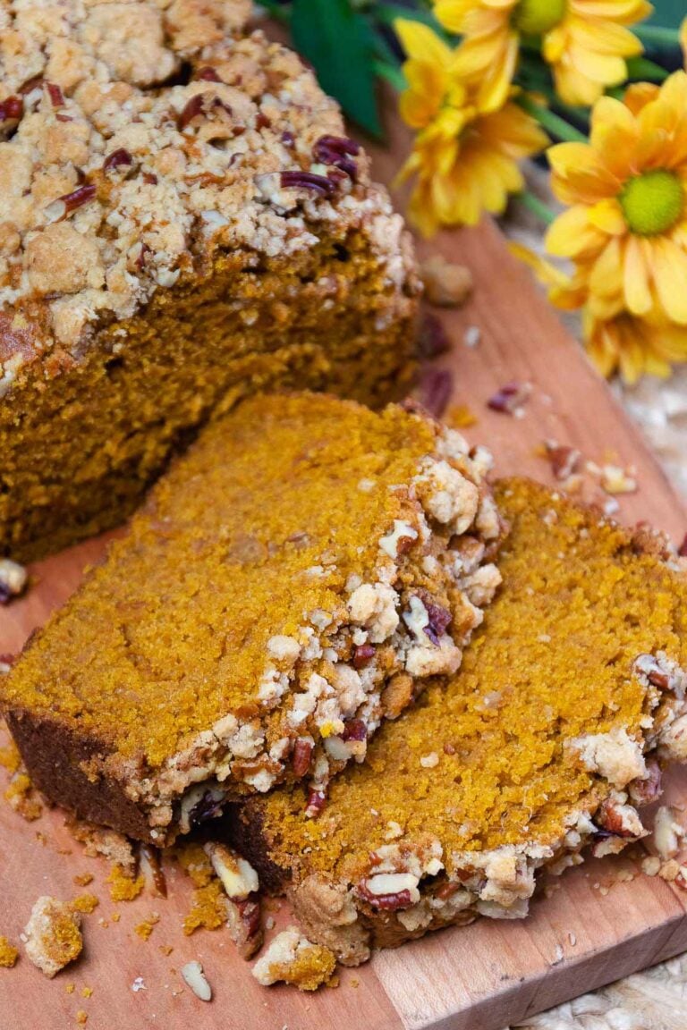 Sliced pumpkin pecan quick bread with crumb topping on wooden board beside yellow flowers
