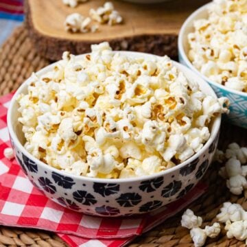 Perfectly Popped Stove Top Popcorn served in patterned bowls on red gingham napkin with loose kernels scattered on woven placemat