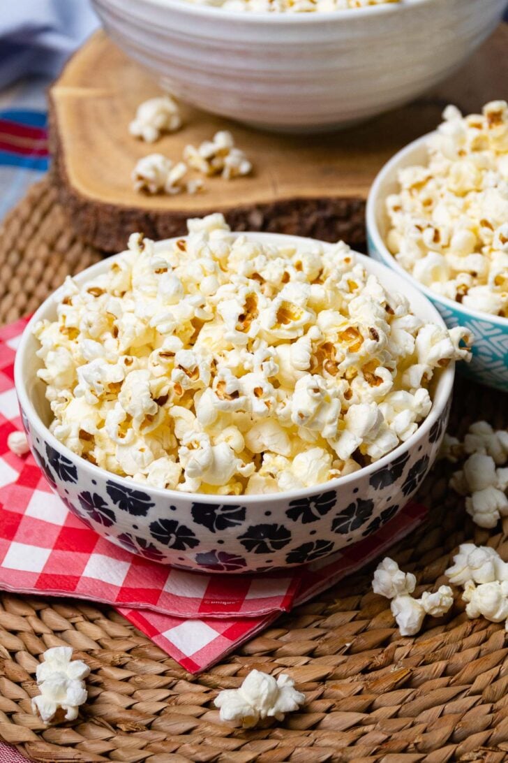 Perfectly Popped Stove Top Popcorn served in patterned bowls on red gingham napkin with loose kernels scattered on woven placemat