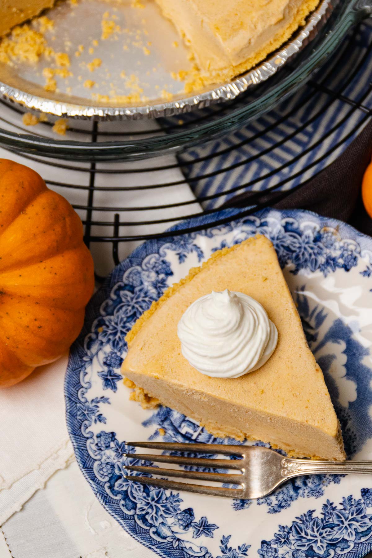 Creamy Pumpkin Icebox Pie slice on decorative blue and white plate with whipped cream dollop, fork, fresh oranges, and pie tin visible