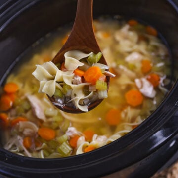 Wooden spoon lifting turkey noodle soup from slow cooker with carrots, celery, and bow-tie pasta in broth