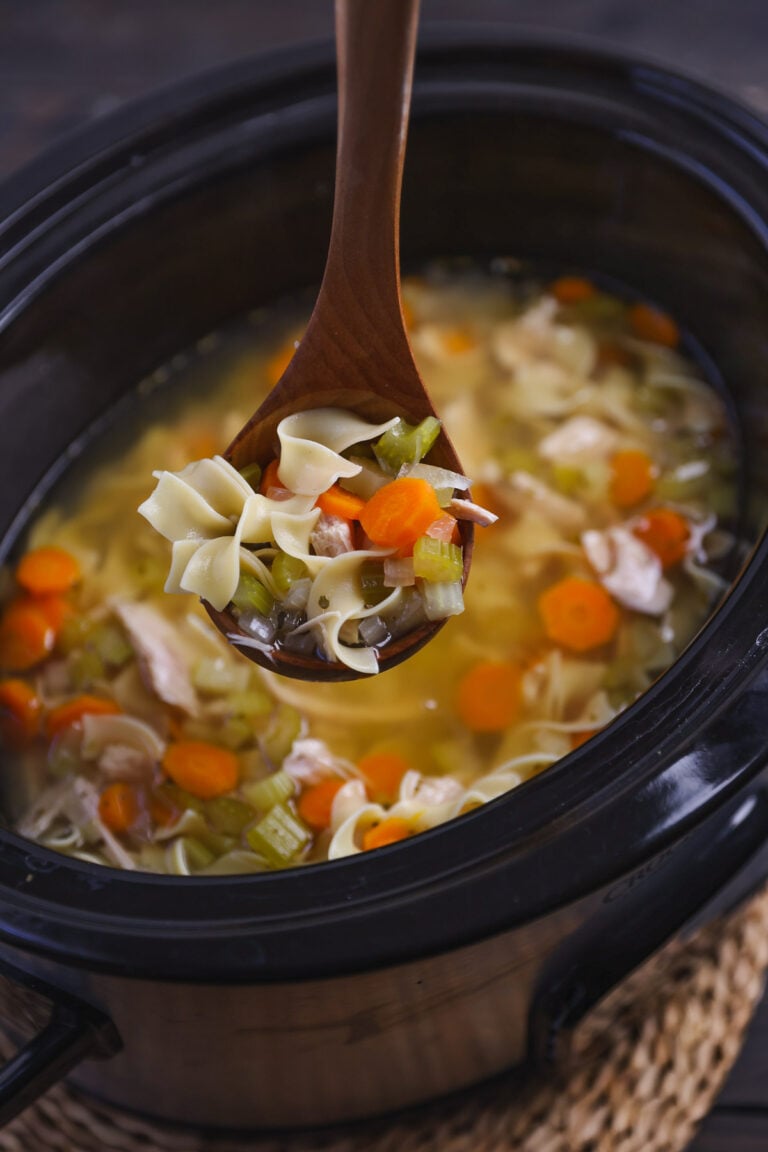Wooden spoon lifting turkey noodle soup from slow cooker with carrots, celery, and bow-tie pasta in broth