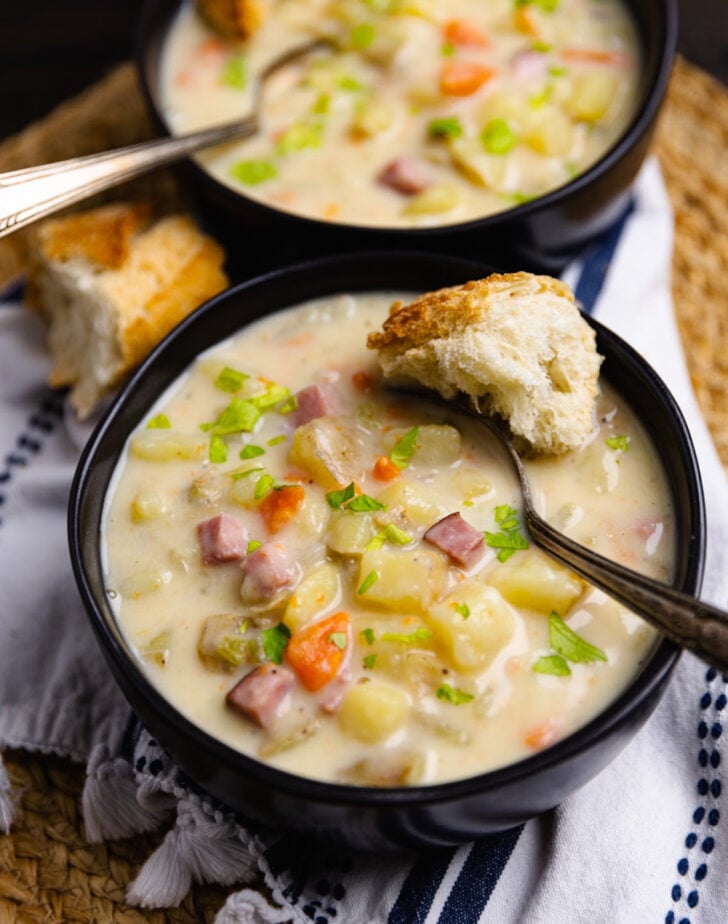 Crock Pot Ham And Potato Soup in black bowls with crusty bread, garnished with green onions and diced ham