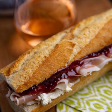 Crusty baguette sandwich filled with sliced ham, creamy spread, and dark red jam on wooden board with wine glass