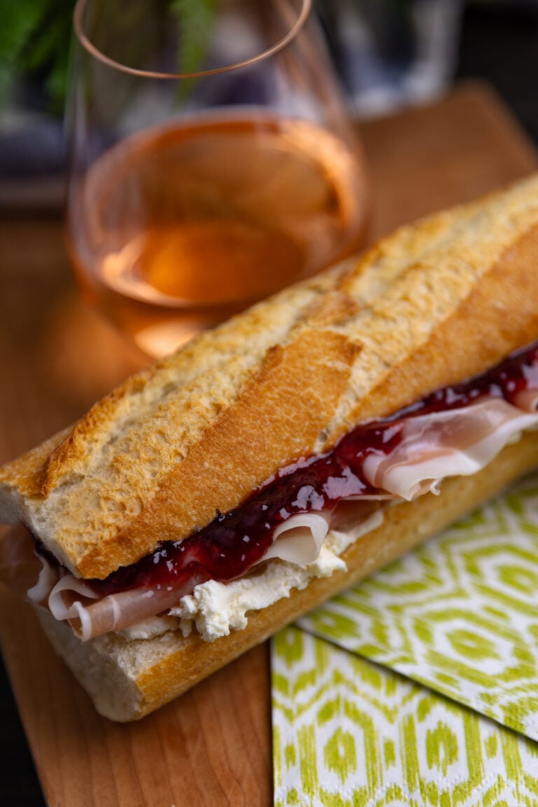 Crusty baguette sandwich filled with sliced ham, creamy spread, and dark red jam on wooden board with wine glass