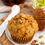 Golden morning glory muffins 6 topped with chopped nuts in brown paper liners on white plate with wooden spoon