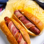 Two air fryer bacon wrapped hot dogs on white plate with yellow bananas and side dishes visible, colorful striped cloth below
