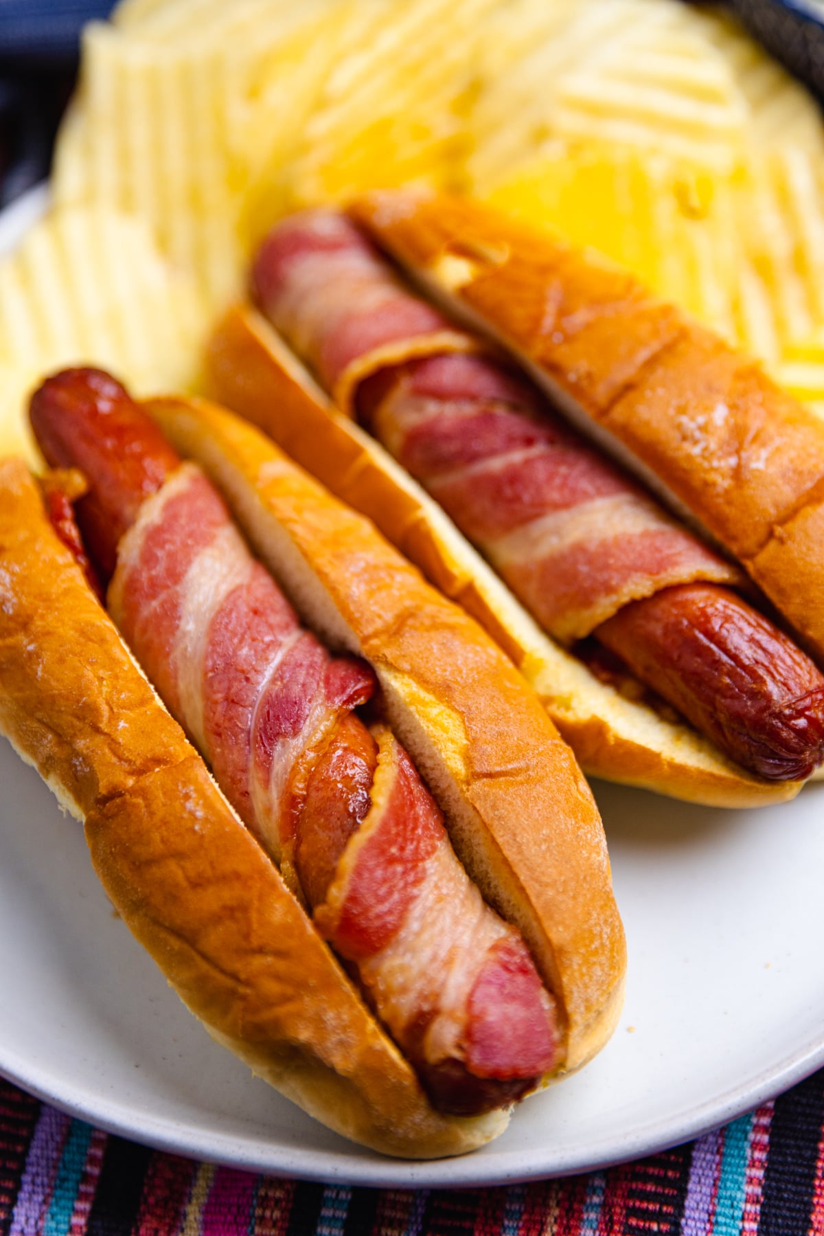 Air Fryer Bacon Wrapped Hot Dogs served on a white plate with toasted buns and shredded cheese garnish, overhead view