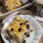 Slice of blueberry buttermilk cake with sugared top on white plate with fork, served with coffee