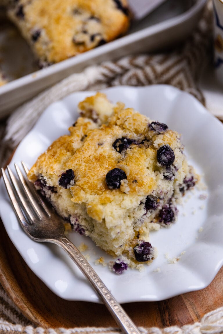 Square slice of Blueberry Buttermilk Cake on white plate with fork, golden crumb texture studded with dark berries