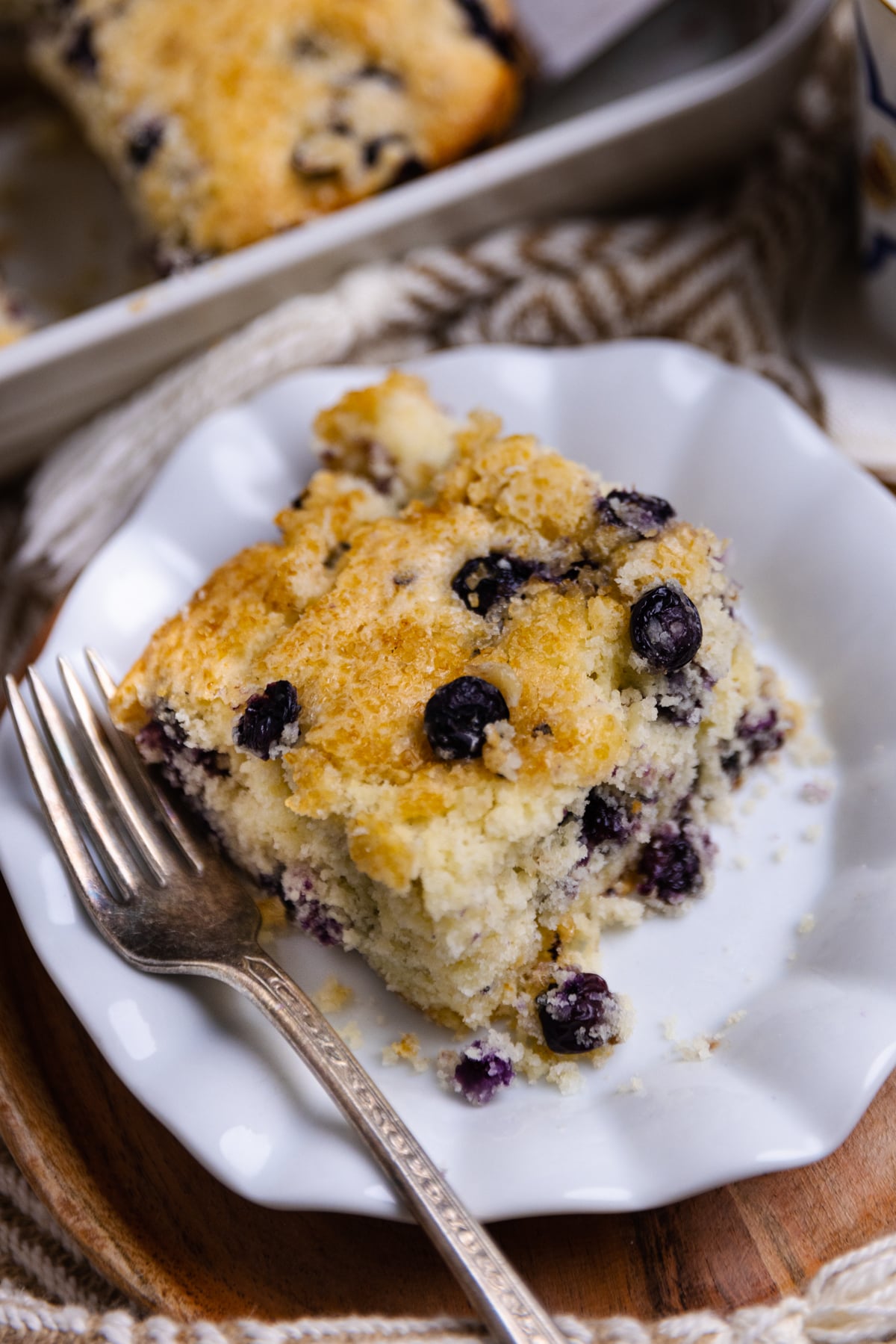 Square slice of Blueberry Buttermilk Cake on white plate with fork, golden crumb texture studded with dark berries