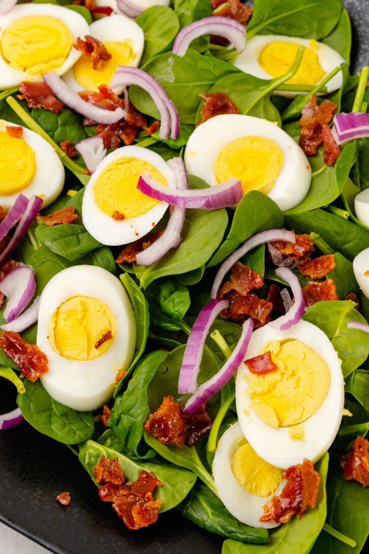 Fresh spinach, sliced boiled eggs, sliced red onion, crispy cooked bacon prepped on a platter for spinach salad.
