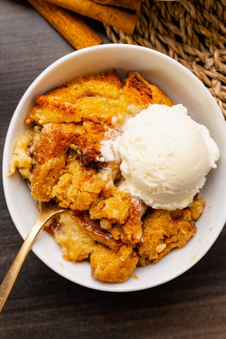 Easy Bread Pudding Recipe served in white bowl with vanilla ice cream scoop and spoon on dark surface