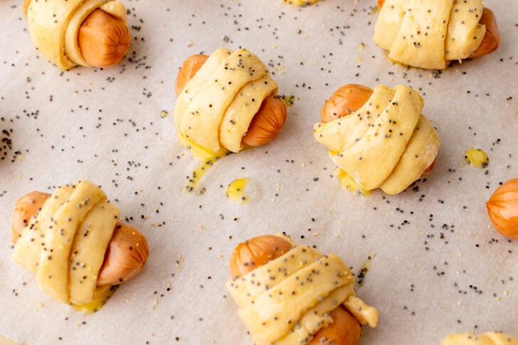 Crescent rolls wrapped around little smokie sausages topped with an egg wash and poppy seeds on a baking sheet ready to cook.