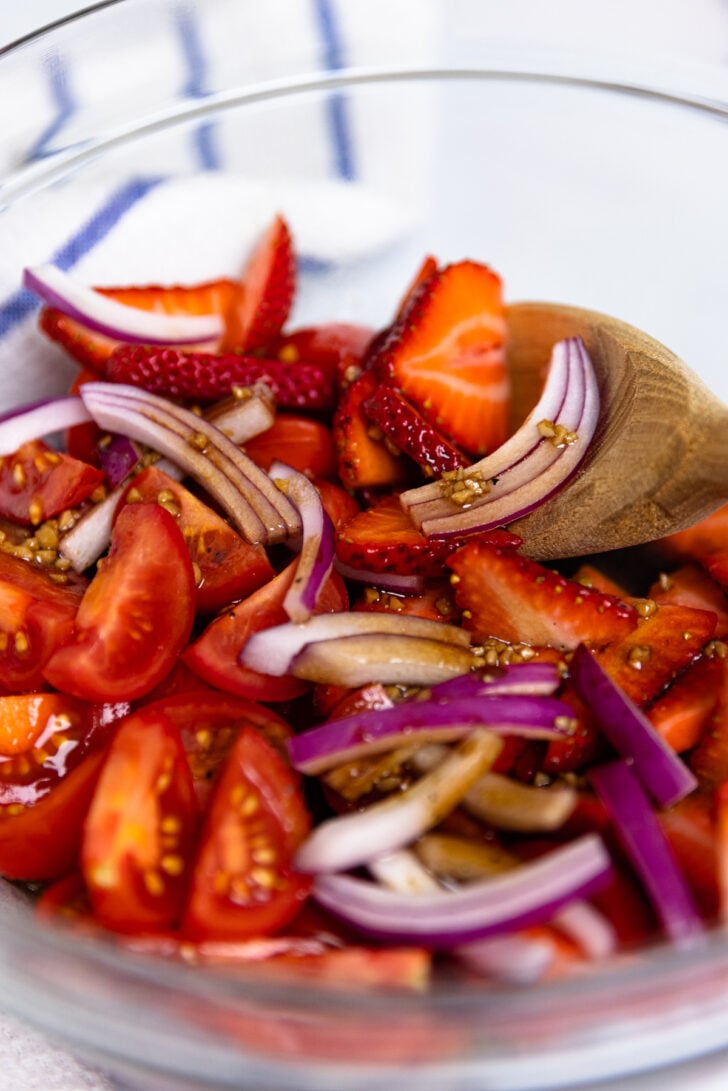 Tomatoes and strawberries with sliced red onion drizzled with balsamic dressing with garlic in a mixing bowl.