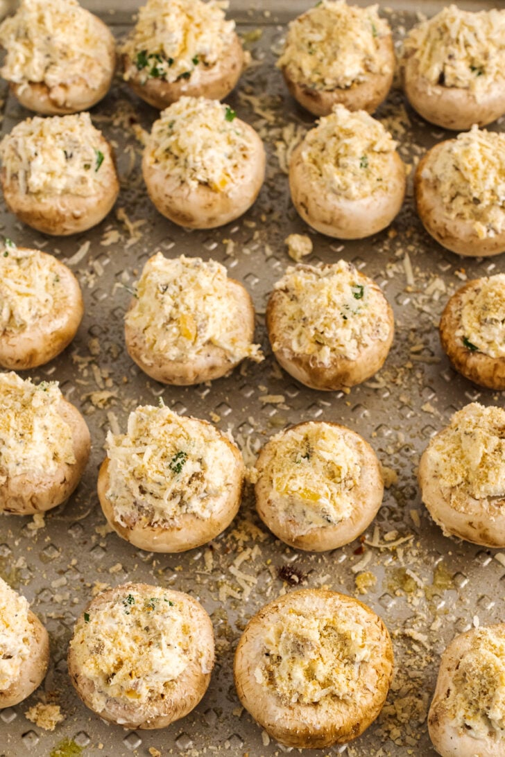 White button mushrooms stuffed with cream cheese, breadcrumbs, parmesan cheese along with garlic and herbs are lined up on a sheet pan ready to be baked.