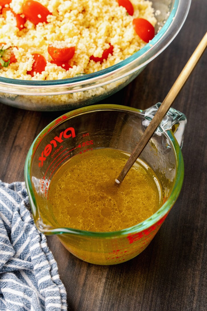 Lemon and olive oil dressing in a mixing cup to be poured over perfectly cooked couscous with fresh tomatoes and basil.