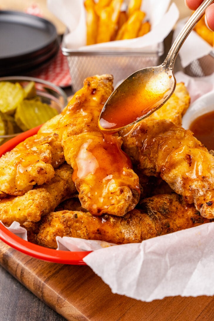 Spoon full of hot honey drizzled over air fryer crispy chicken tenders served Nashville style along with pcikles and french fries.