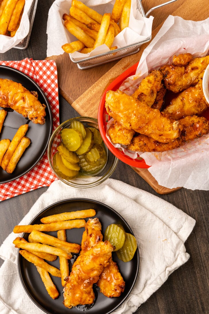 Hot chicken tenders, Nashville style served with pickles and french fries on a black plate for dinner, a snack or for lunch.
