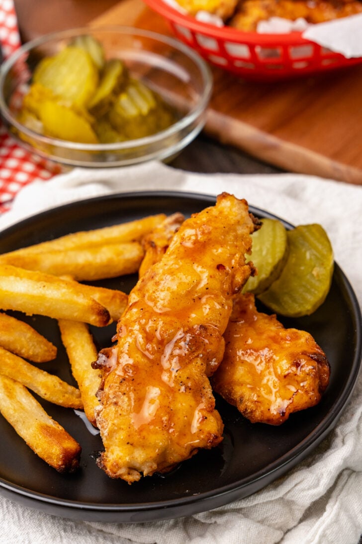 Hot chicken tenders, Nashville style served with pickles and french fries on a black plate for dinner.