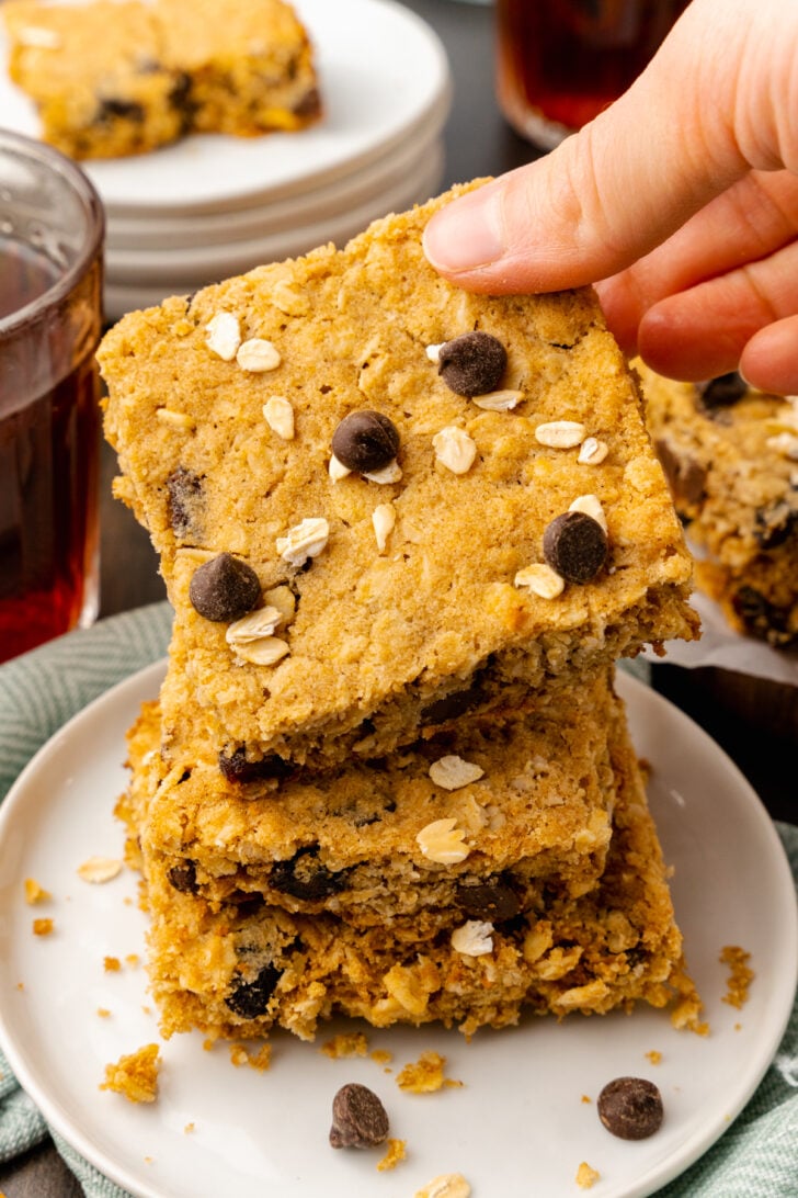 Hand holding oatmeal chocolate chip cookie bar with chocolate chips on the top served with a cup of hot tea for an afternoon snack.