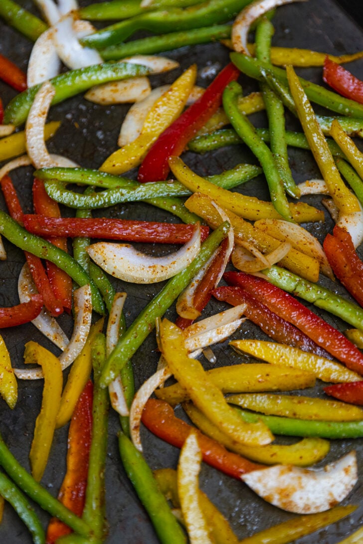 Red, yellow, and green bell peppers sliced along with onion tossed in fajita seasoning on a sheet pan to be roasted for a shrimp dinner.