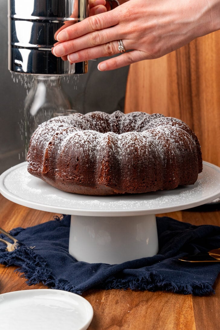 Hands holding a sifter full of powdered sugar over a Southern chocolate pound cake sitting on a white cake stand.