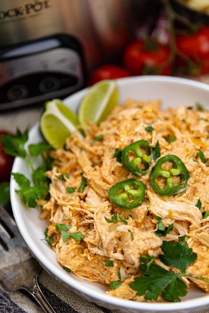 White bowl of shredded cream cheese salsa chicken cooked in the crock pot topped with jalapenos, fresh parsley and lime wedges.