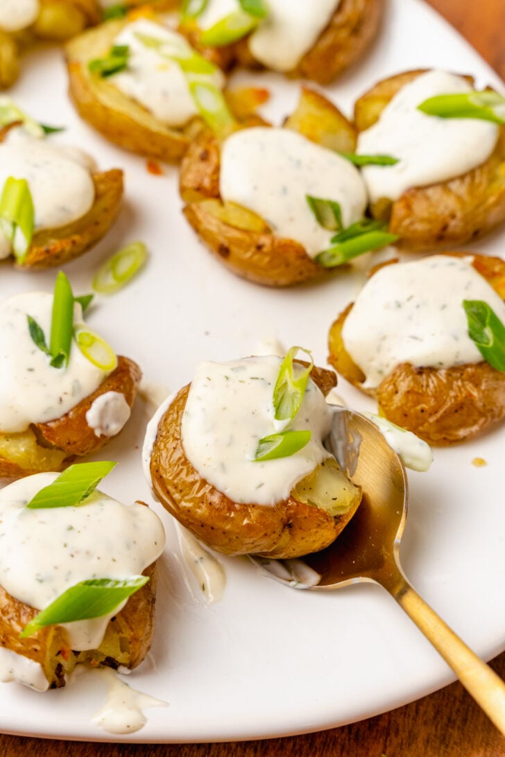 Crushed baby potatoes topped with homemade ranch dressing and chopped green onions served on a white platter and a gold spoon.