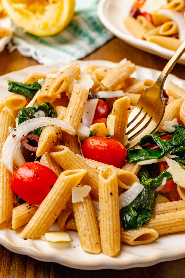 Whole wheat penne pasta tossed with fresh tomatoes, spinach, black olives in lemon juice and olive oil the topped with shaved Parmesan Cheese served on a white plate with a gold fork for lunch.