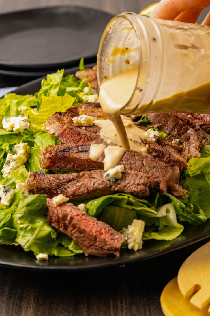 Creamy vinaigrette salad dressing being poured over grilled ribeye steak, lettuce and blue cheese for a hearty dinner salad.