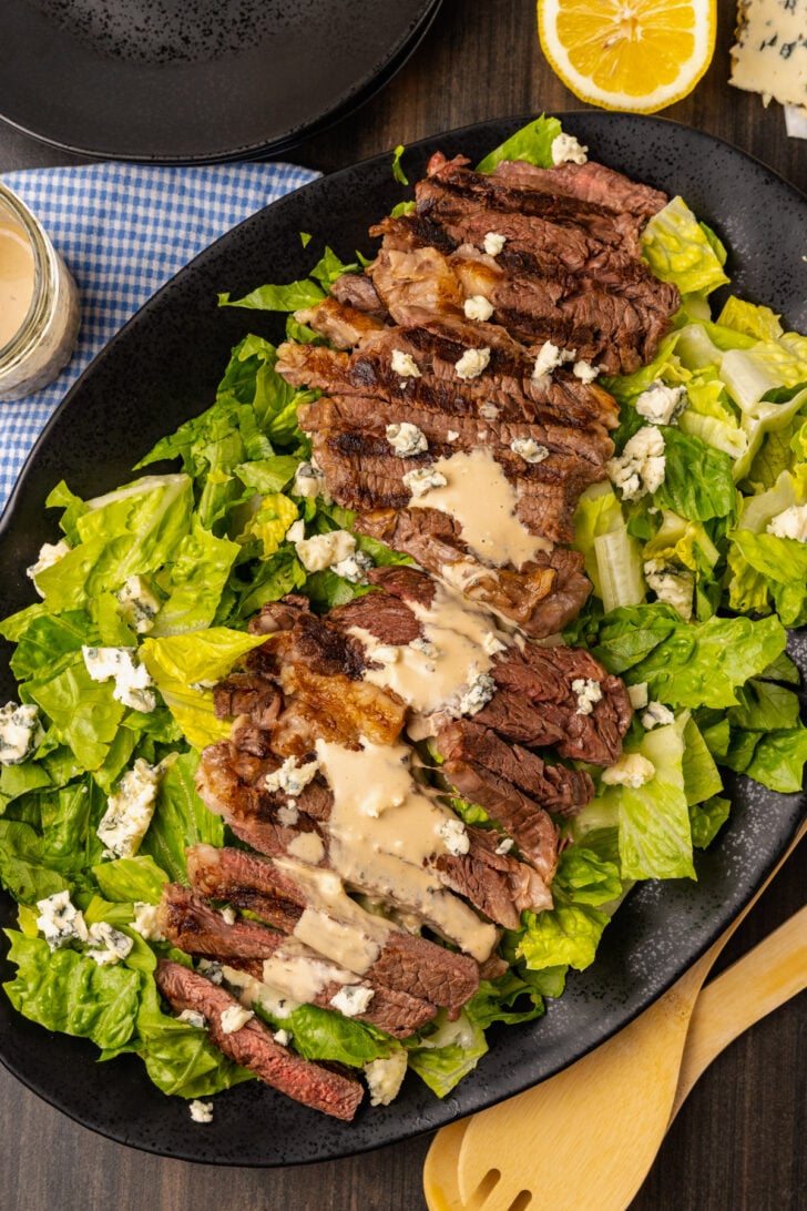 Grilled Steak Salad with blue cheese, creamy vinaigrette salad dressing and chopped romaine lettuce with blue cheese crumbles served on a black platter.