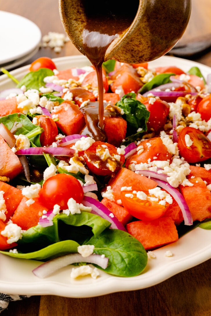 Homemade balsamic dressing being drizzled over watermelon, spinach, red onion and feta salad.