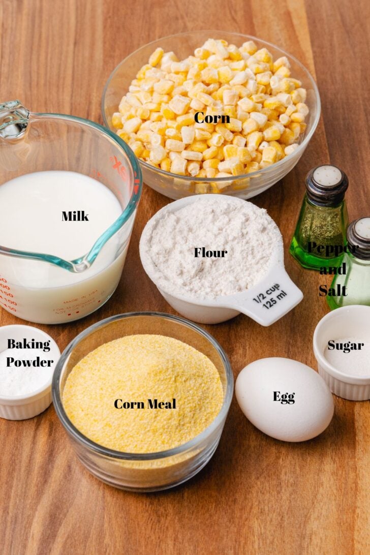 Corn, milk, salt, pepper, sugar, egg, corn meal, baking powder, and flour ingredients on a brown board.
