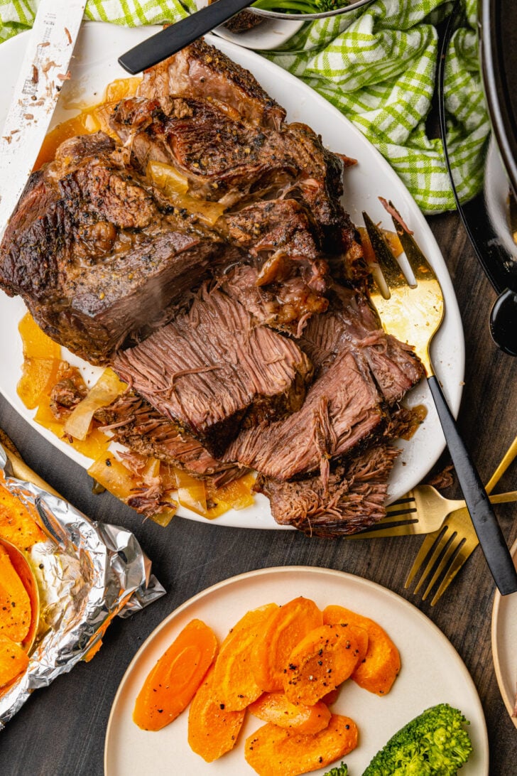Sliced beef pot roast cooked with onions on a white platter served with carrots and steamed broccoli.