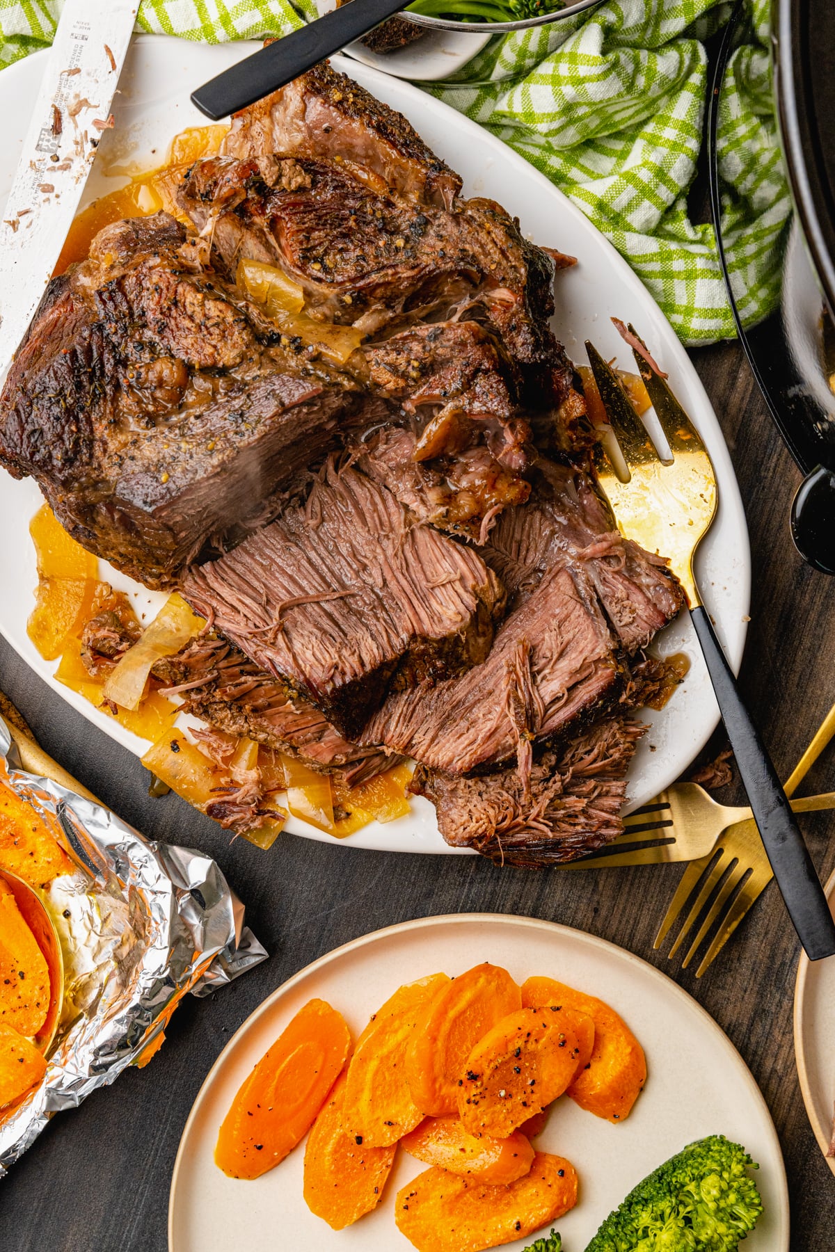 Sliced beef pot roast cooked with onions on a white platter served with carrots and steamed broccoli.