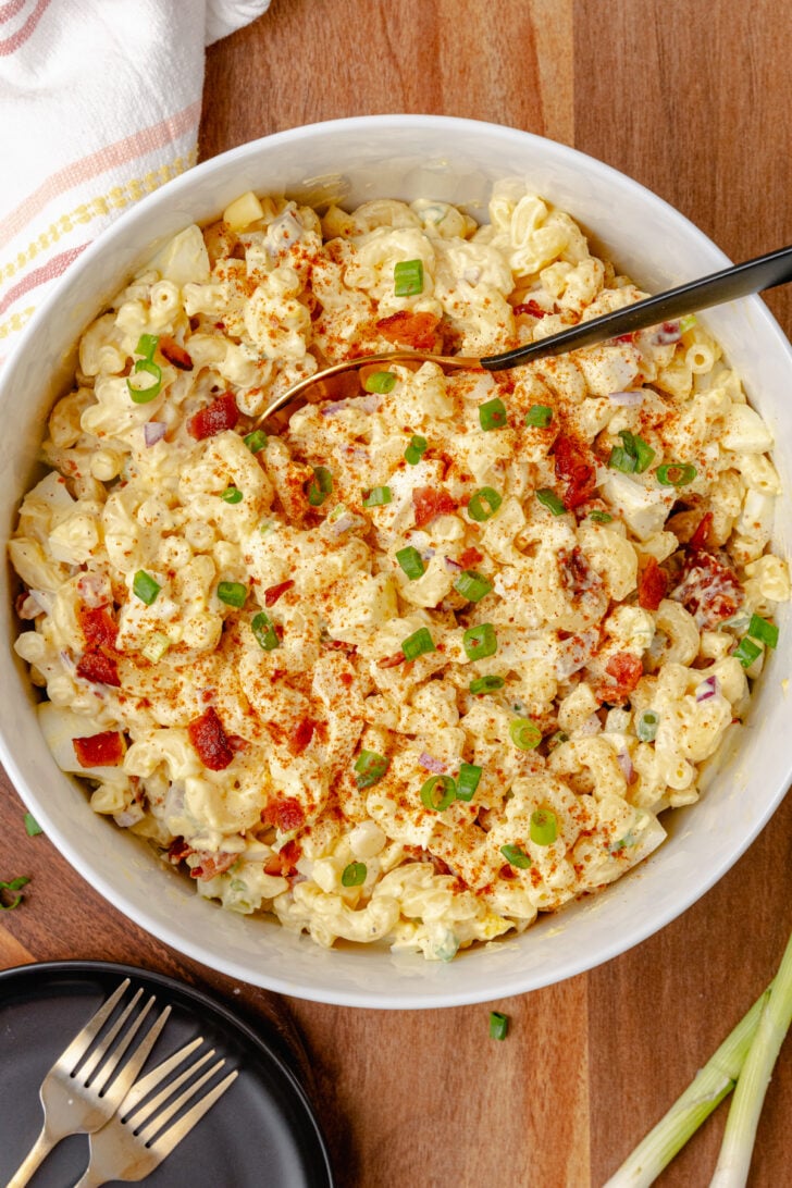 Creamy Deviled Egg Pasta Salad in white bowl with elbow pasta, diced red pimentos, green onions, paprika garnish, and serving spoon on woode