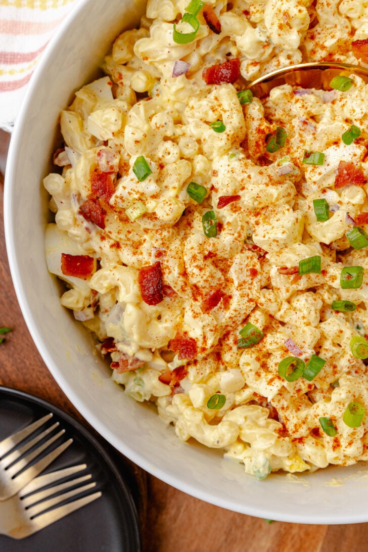 Creamy and full of flavor, Deviled Egg Pasta Salad is a side dish classic that perfect for any family gathering, cookout, or potluck. Made with elbow macaroni, boiled eggs, celery, bacon and topped with green onions all in a delicious mayonnaise based dressing topped with a sprinkle of paprika served in a white bowl.