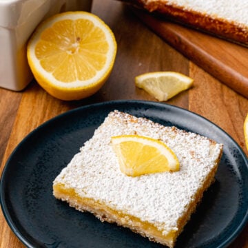 A lemon bar topped with a lemon slice served on a black plate for dessert.