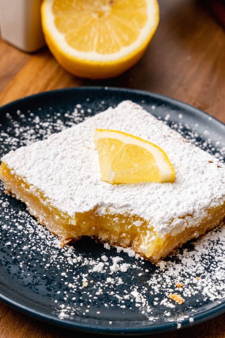 Lemon square topped with powdered sugar and a lemon slice on a black plate.