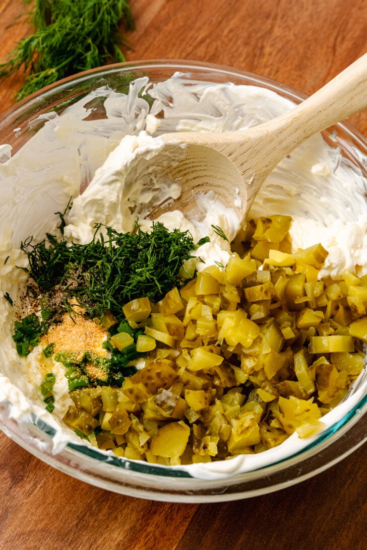 Glass bowl with cream cheese, chopped dill pickles, fresh dill, and garlic powder stirred with a wooden spoon to make dill pickle dip.