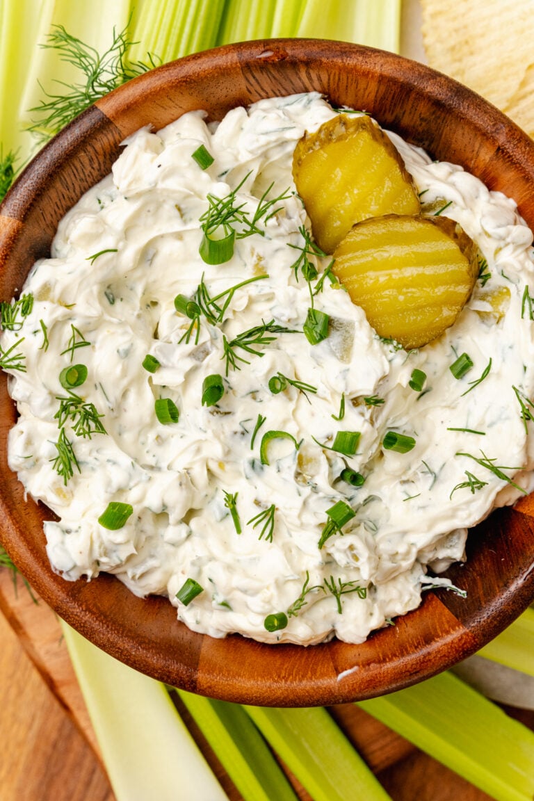 Homemade dill pickle dip served in a wooden bowl with fresh dill and chives garnish along with celery sticks and potato chips.