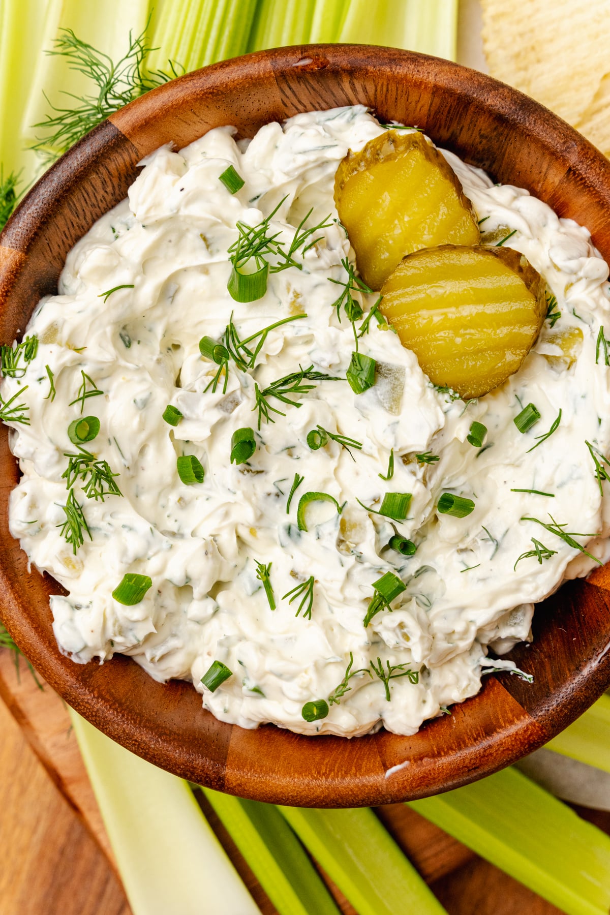 Homemade dill pickle dip served in a wooden bowl with fresh dill and chives garnish along with celery sticks and potato chips.