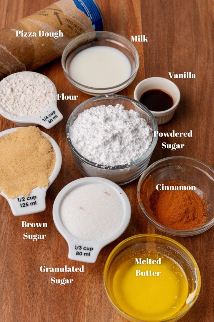 Pizza dough, milk, vanilla, powdered sugar, cinnamon, melted butter, granulated sugar, brown sugar, flour in bowls on a wooden board.