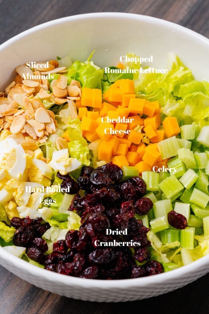 Chopped lettuce, cubed cheddar cheese, chopped celery, chopped hard boiled eggs, sliced almonds and dried cranberries in a white bowl sitting on a wooden cutting board.