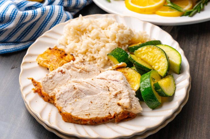Sliced chicken breasts cooked in the air fryer served with sautéed zucchini and rice on a white plate on a wooden table.
